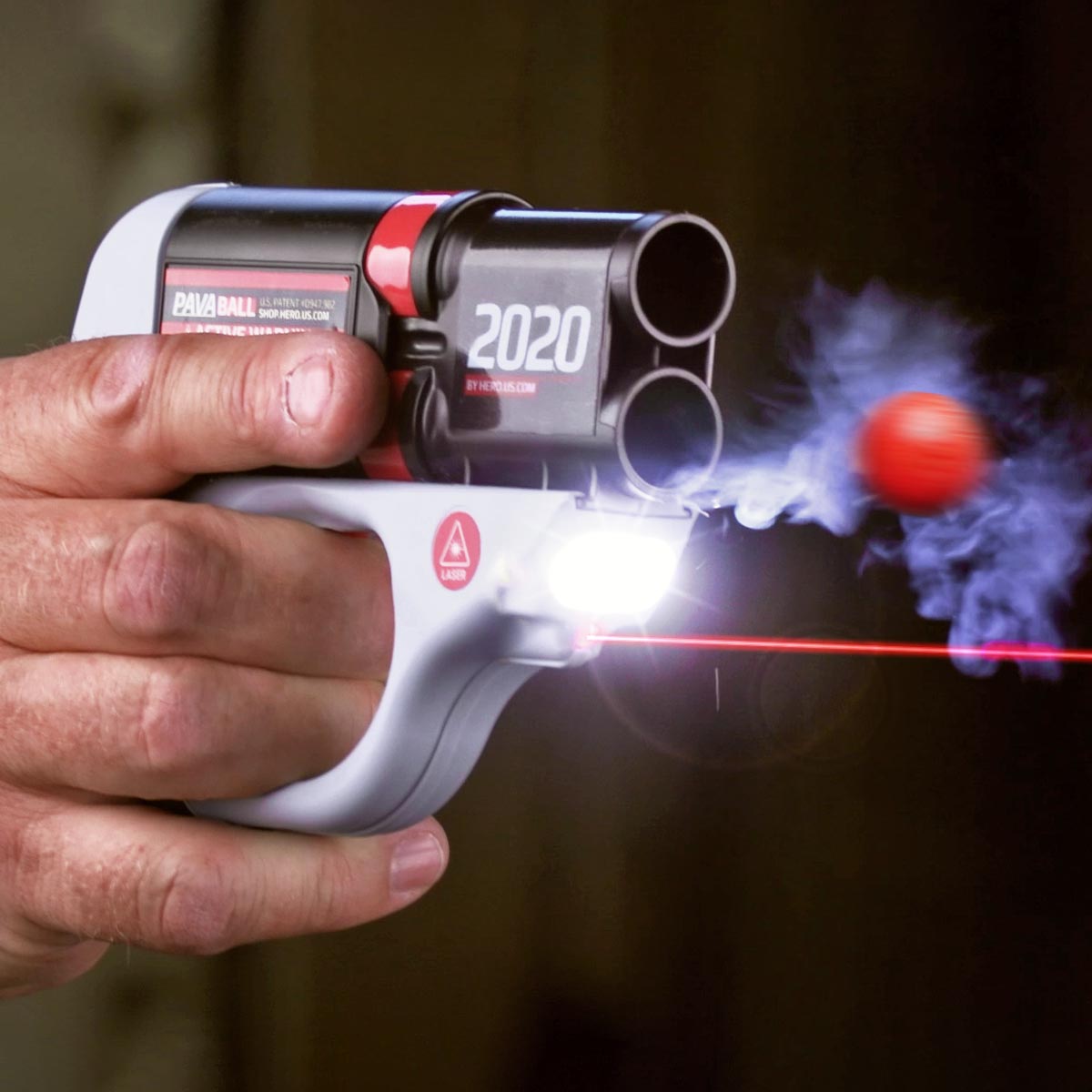 A hand squeezes the trigger, firing HERO 2020. A projectile flies from the bottom barrel. A laser and bright light are lit on the device.